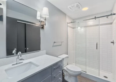 Modern bathroom with white fixtures, glass-enclosed shower, wall-mounted mirror, and vanity with sink and storage drawers at Merchants Plaza.