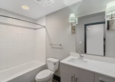 Modern bathroom with bathtub, toilet, and vanity with sink and mirror. White tiles and walls, and two wall-mounted lamps at Merchants Plaza.