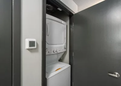 Stacked washer and dryer inside a closet with partially open dark doors; a thermostat is on the left wall at Merchants Plaza.