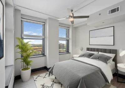 A modern bedroom with a double bed, two windows, a ceiling fan, a plant, and minimalist decor in shades of gray and white at Merchants Plaza.