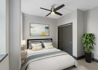 A modern bedroom with a bed, artwork on the wall, a ceiling fan, and a potted plant by the closet door at Merchants Plaza.