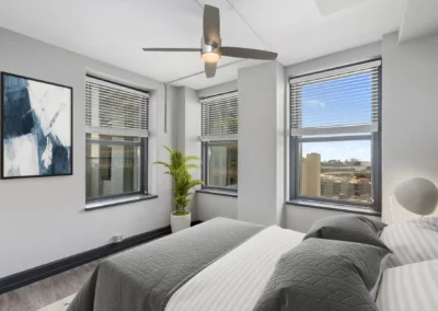 Modern bedroom with large windows, a ceiling fan, and minimalistic decor. A plant and abstract painting add accents at Merchants Plaza.
