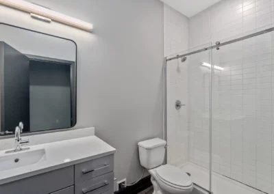 Modern bathroom with a vanity, wall mirror, toilet, and a glass-enclosed shower area with white tiled walls at Merchants Plaza.