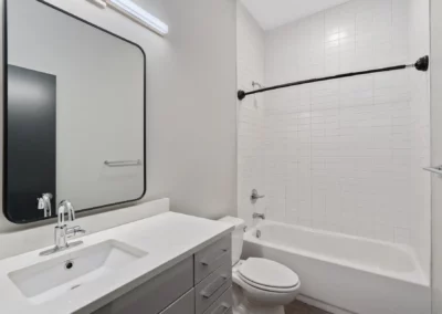 Modern bathroom with a white vanity, large rectangular mirror, toilet, and bathtub with a tiled wall and shower curtain rod at Merchants Plaza.