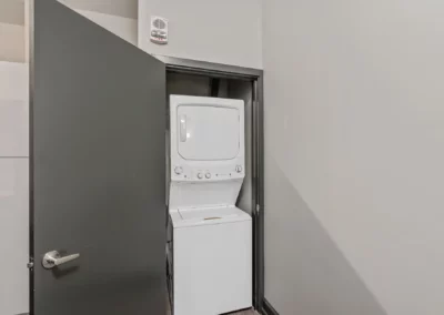 A stacked washer and dryer unit is placed in a small, open closet next to a light gray wall and an open dark gray door at Merchants Plaza.