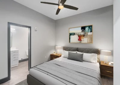 A modern bedroom with a large bed, nightstands, lamps, artwork, ceiling fan, and an open door leading to a bathroom at Merchants Plaza.