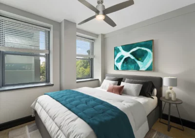 Modern bedroom with a double bed, teal and white bedding, abstract art, large windows, grey walls, and ceiling fan at Merchants Plaza.