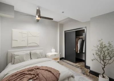 Minimalist bedroom with a bed, nightstand, ceiling fan, abstract wall art, and an open closet next to a potted plant at Merchants Plaza.